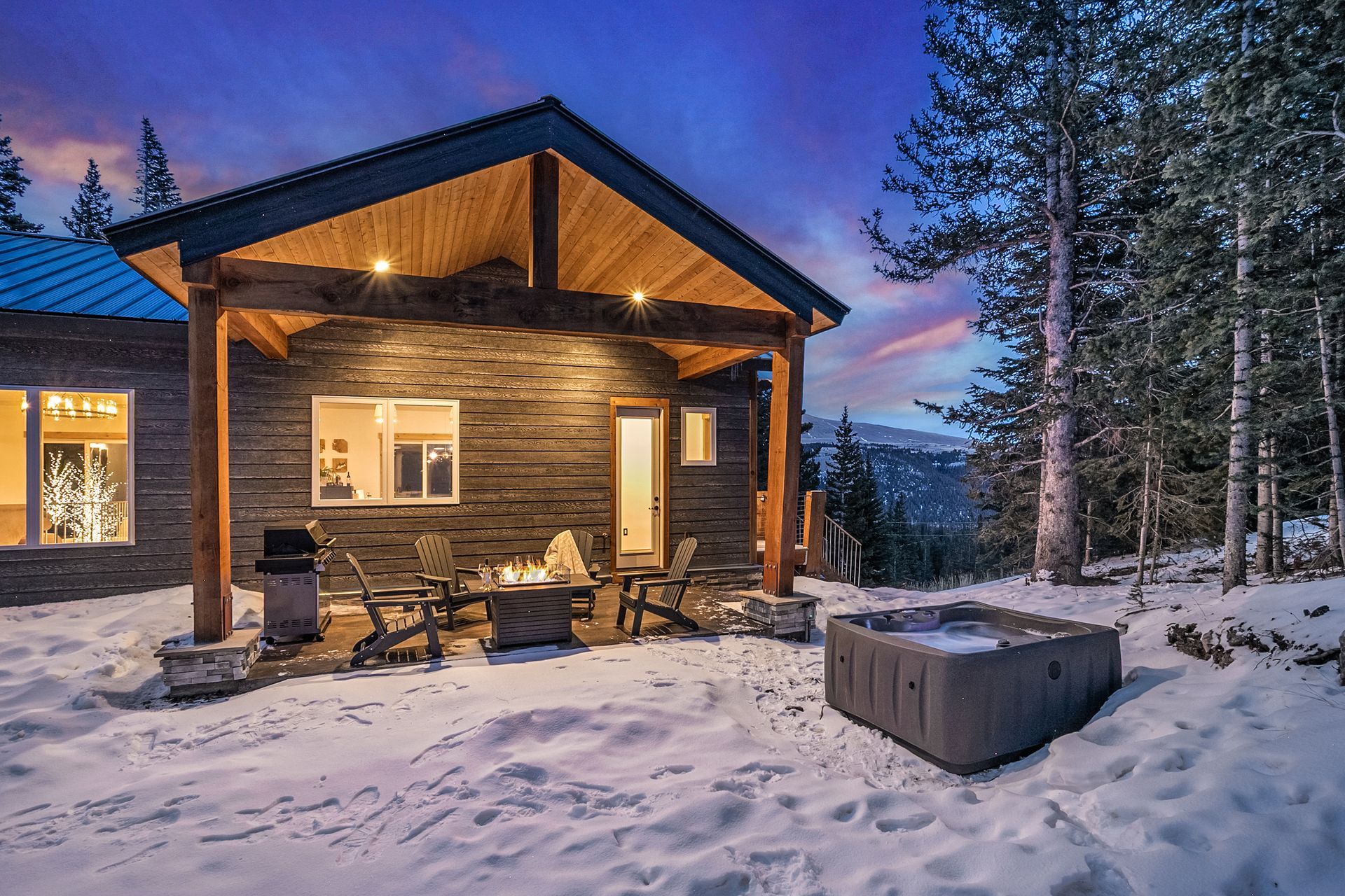 A snowy patio at dusk with a covered wooden porch, stone-tiled walls, outdoor seating, a fire pit, and a small hot tub.
