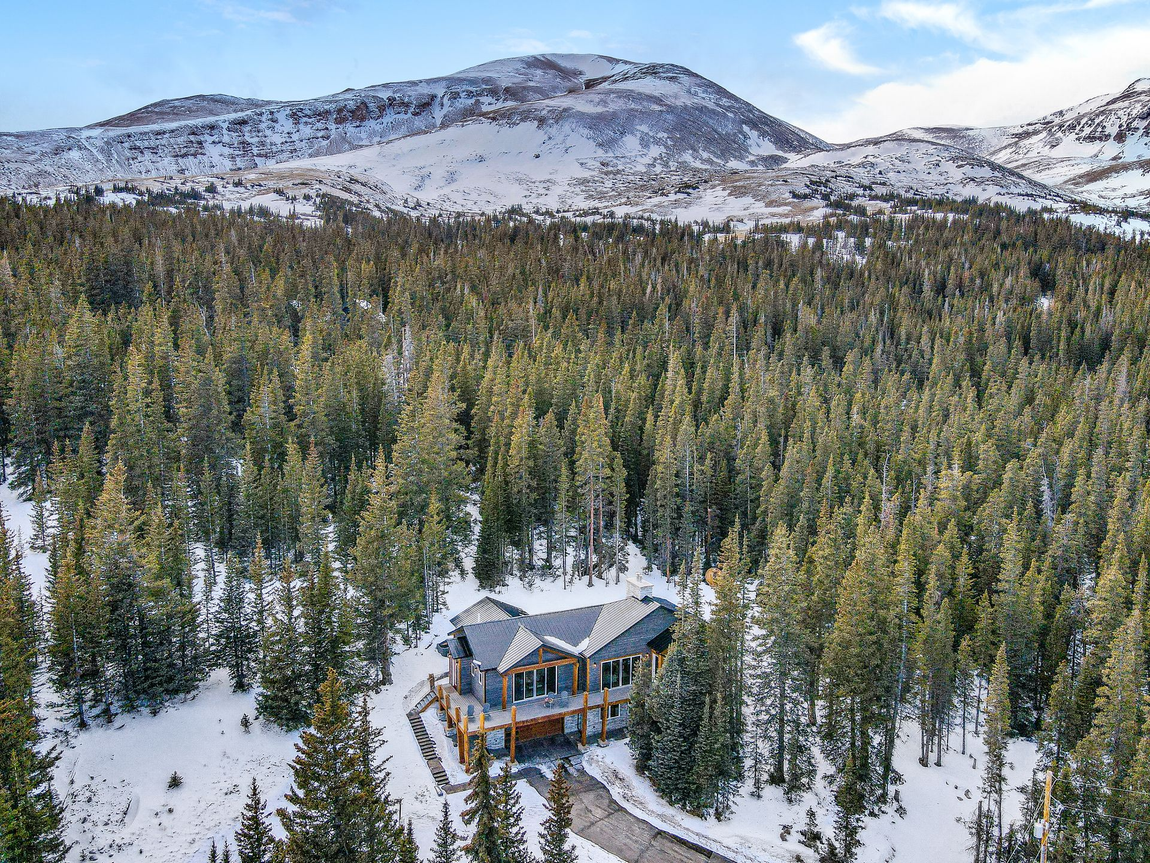Snowy cabin nestled in a pine forest below mountains under a blue sky