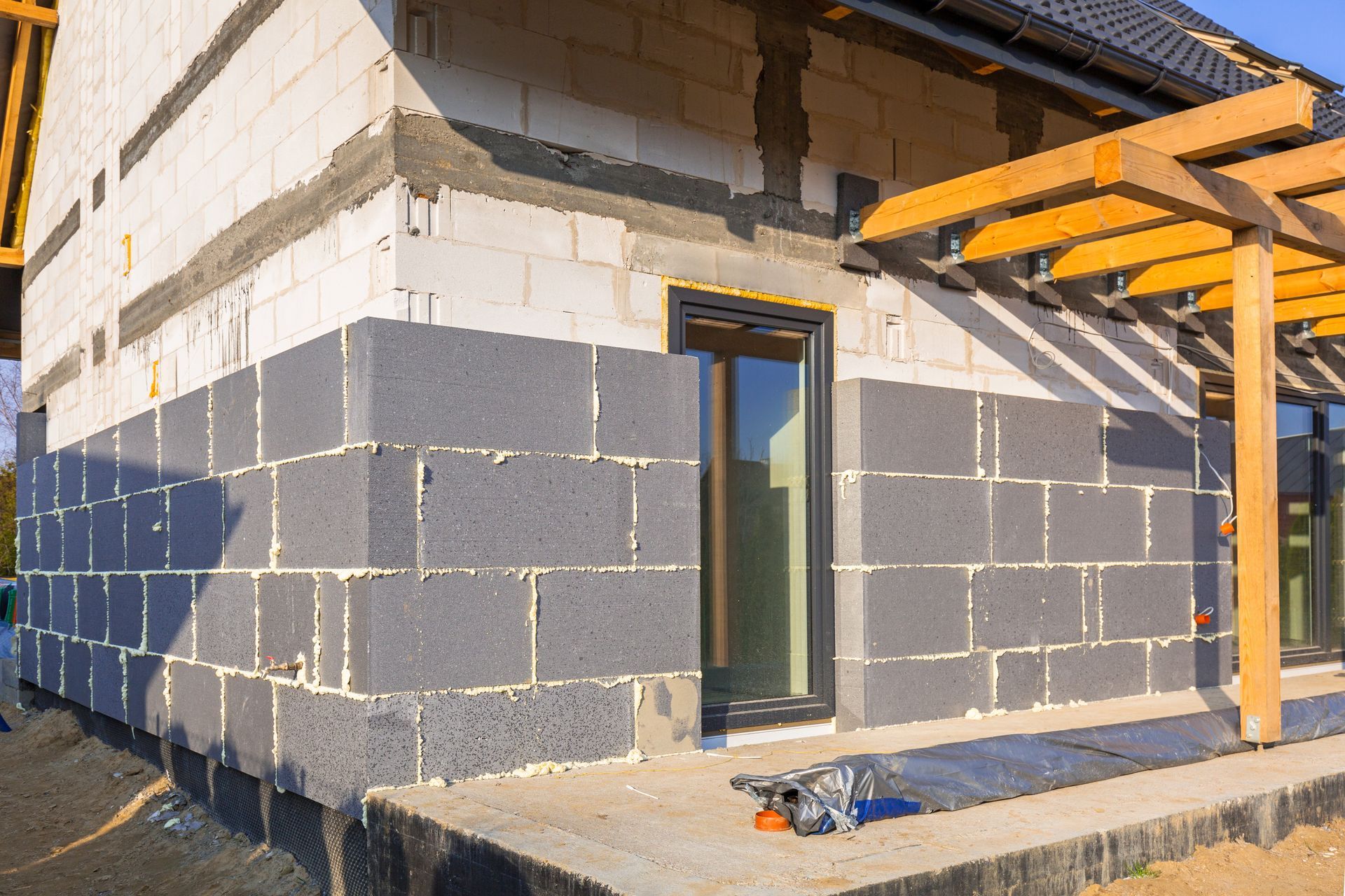 Extérieur d'une maison en construction ; murs en parpaings, fenêtre et pergola en bois.