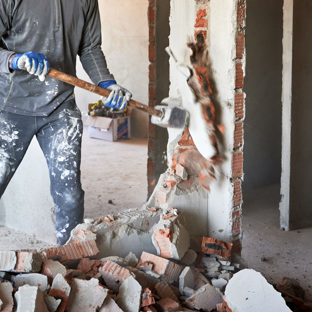 Une personne démolit un mur de briques à coups de masse à l'intérieur d'un bâtiment, entourée de débris.
