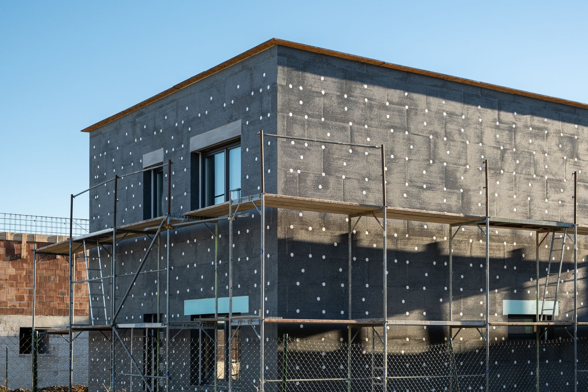 Bâtiment en construction, panneaux d'isolation gris, échafaudages, fenêtres, ciel bleu.