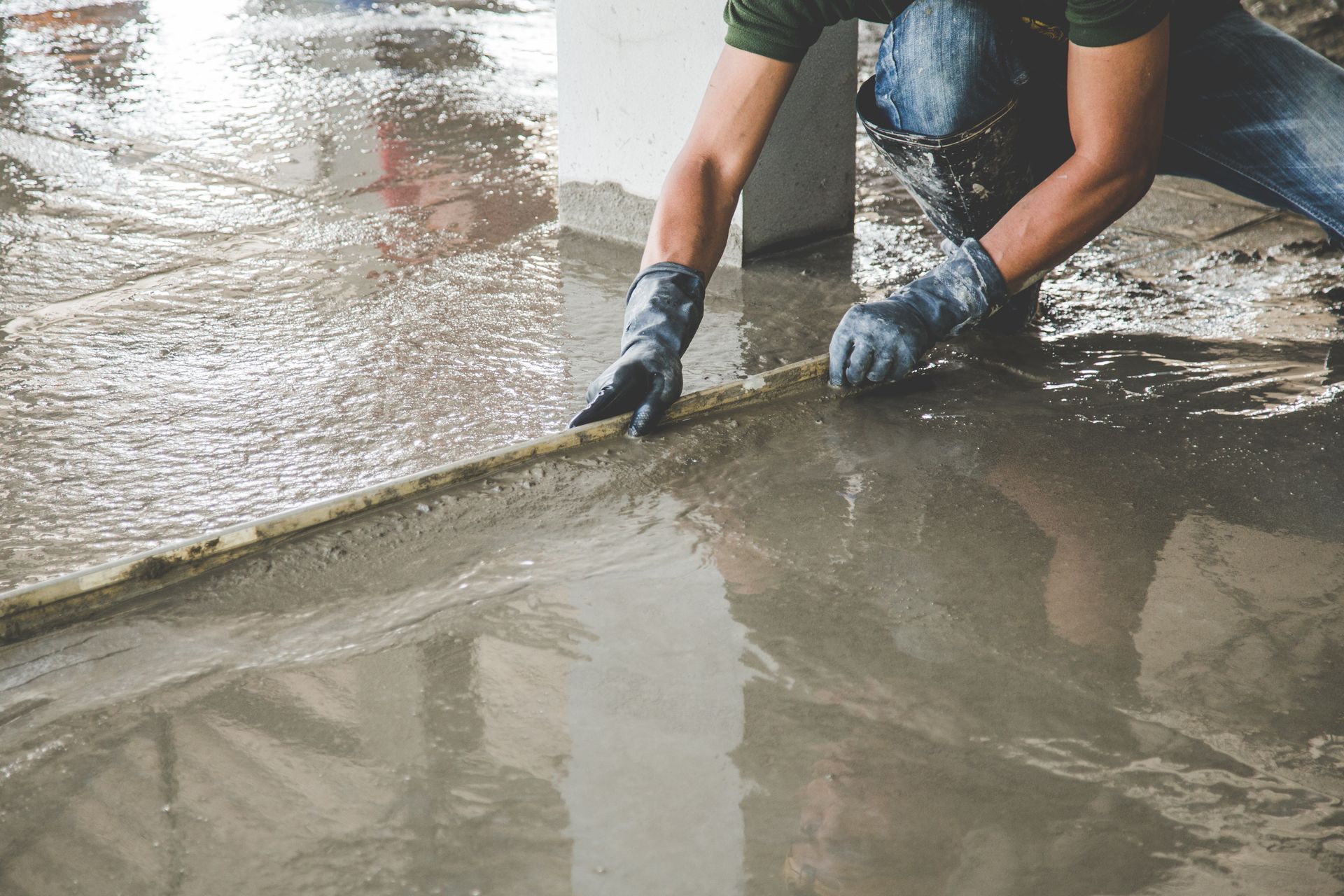 Une personne agenouillée lisse du béton frais avec des gants. Chantier aux surfaces grises et humides.