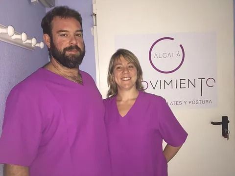 Dos personas con uniformes morados posan frente a un cartel que dice