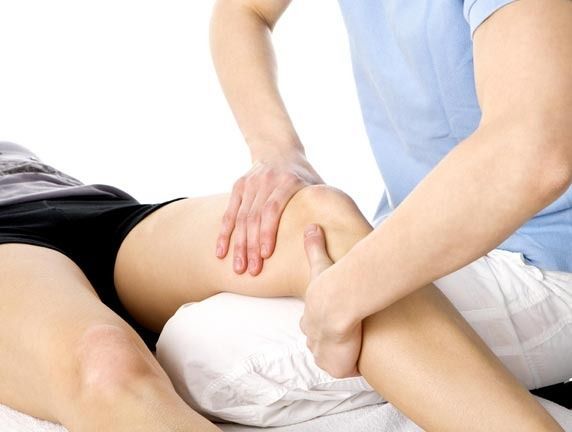 Terapeuta masajeando la pierna y la rodilla de un paciente, probablemente para fisioterapia en una mesa de examen blanca.