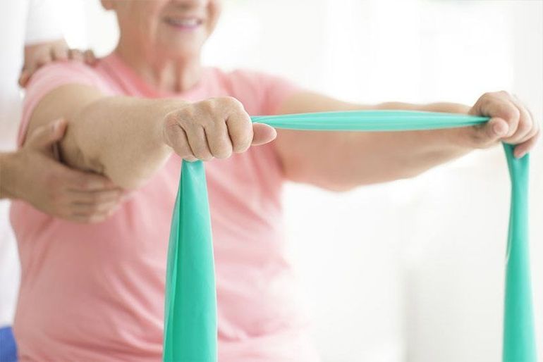 Mujer mayor con camisa rosa usando una banda de resistencia color verde azulado, asistida por las manos de otra persona para fisioterapia.