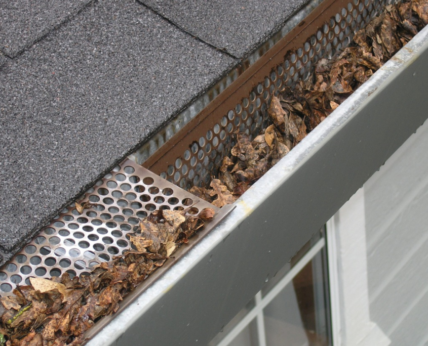 Una canaleta llena de hojas secas y marrones, parcialmente cubierta por una rejilla metálica junto a un tejado de tejas.
