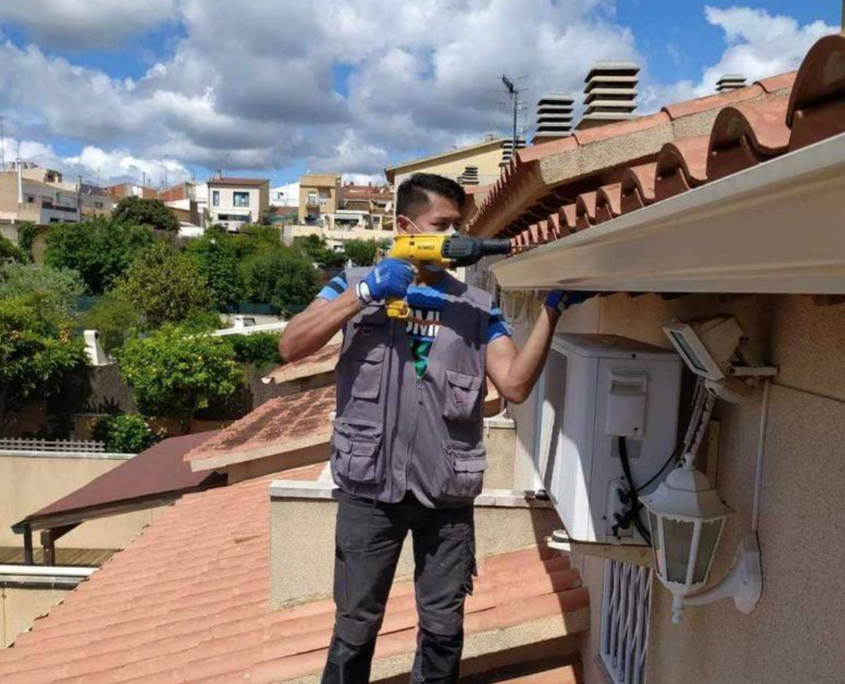 Un trabajador, vestido con un chaleco gris y guantes, utiliza un taladro eléctrico para instalar un canalón de color beige en el tejado de una casa con tejas.