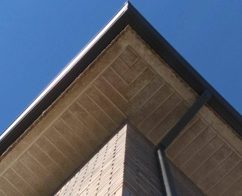 Vista desde abajo de la esquina de una casa de ladrillo, con un alero texturizado de color beige, molduras de color marrón oscuro y un cielo azul despejado.