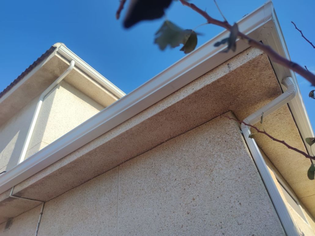 Primer plano del exterior de una casa que muestra paredes de estuco color crema, un sistema de canalones blancos y un alero de techo sobresaliente.