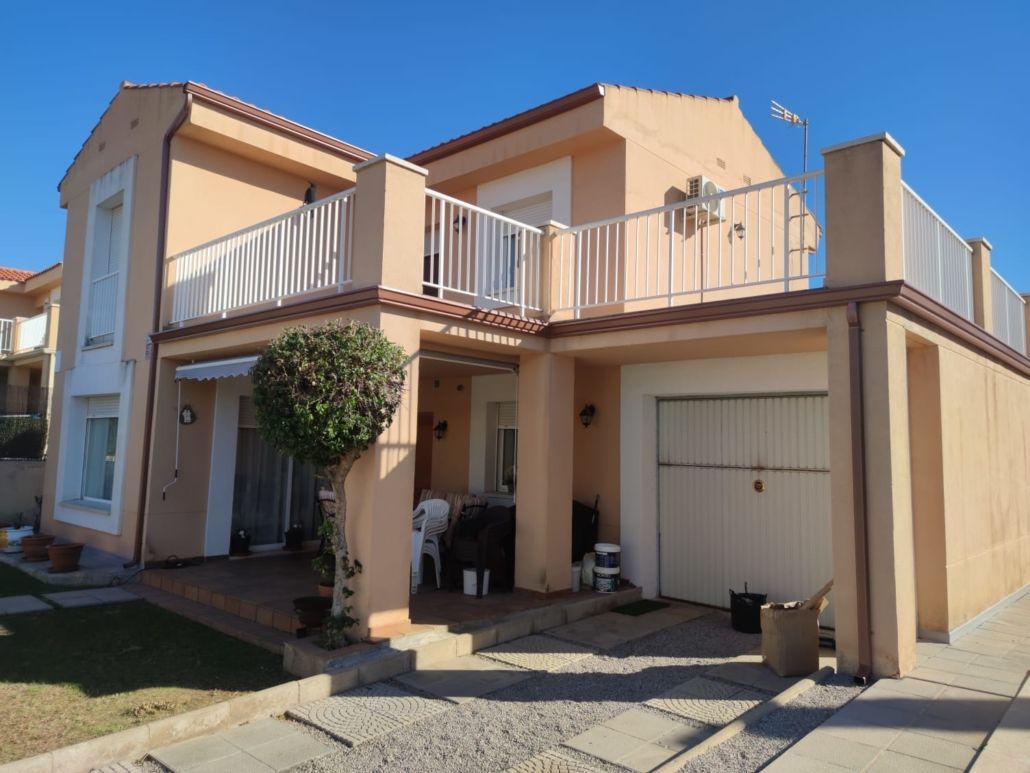 Una casa suburbana de dos plantas, color melocotón, con balcón, patio, garaje y un pequeño árbol bien cuidado bajo un cielo azul despejado.