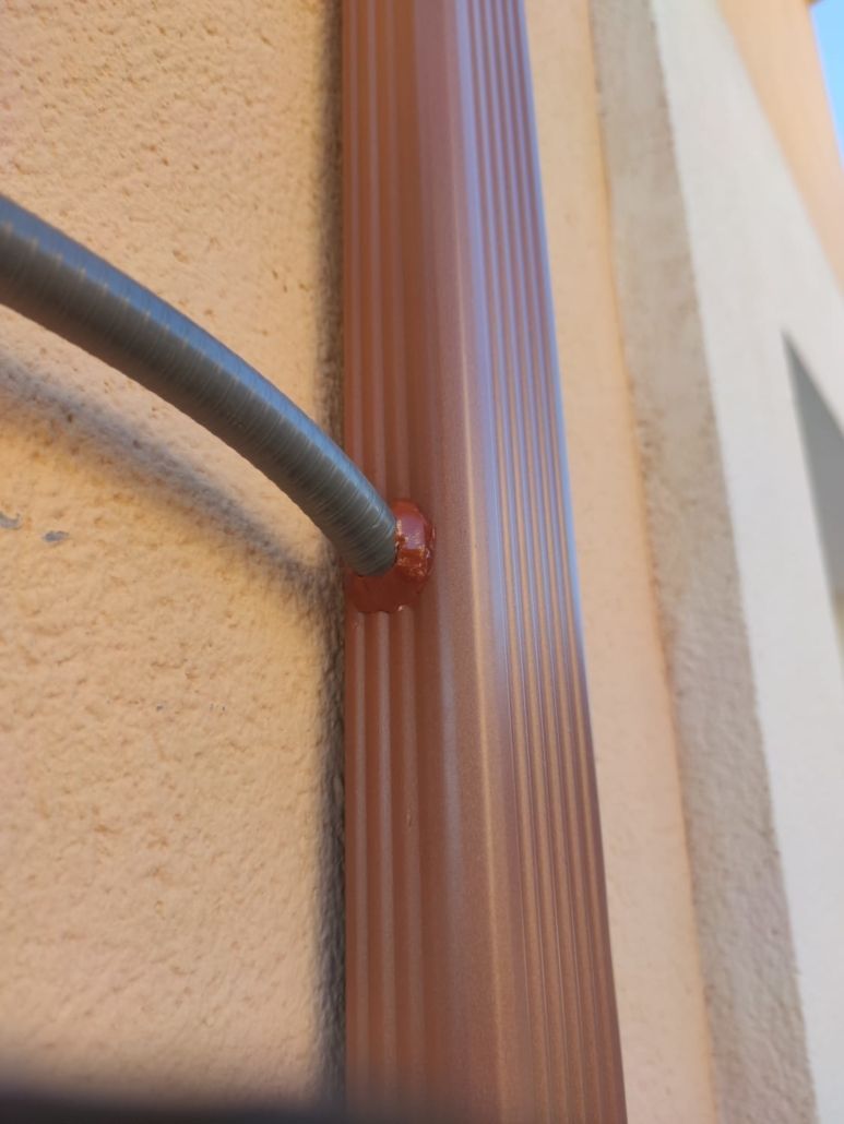 Un cable gris entra en un bajante metálico marrón en una pared de estuco, sellado con un sellador de color marrón rojizo.