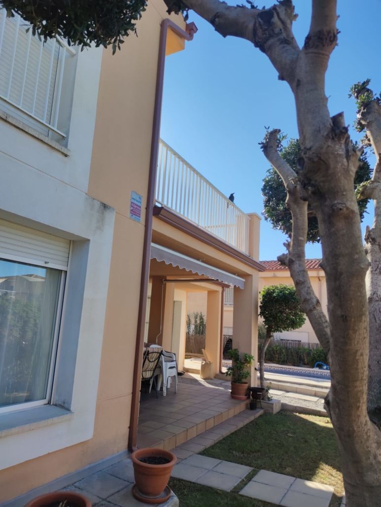 Una casa beige de dos plantas, bañada por el sol, con patio cubierto, balcón, bajante marrón y un gran árbol en el jardín delantero.