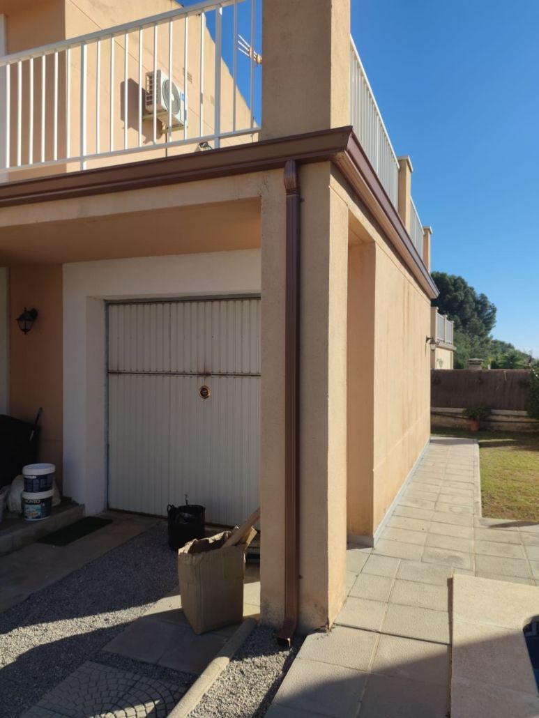 Vista lateral de un edificio de color beige con una puerta de garaje blanca, un bajante marrón, un balcón y un camino pavimentado en un patio soleado.