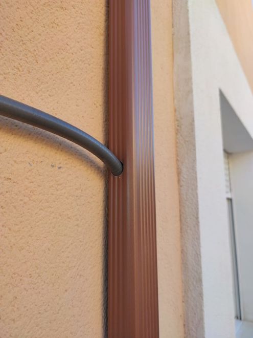 Un cable gris entra por un agujero en un bajante marrón acanalado montado en una pared de estuco junto a un marco de puerta blanco.