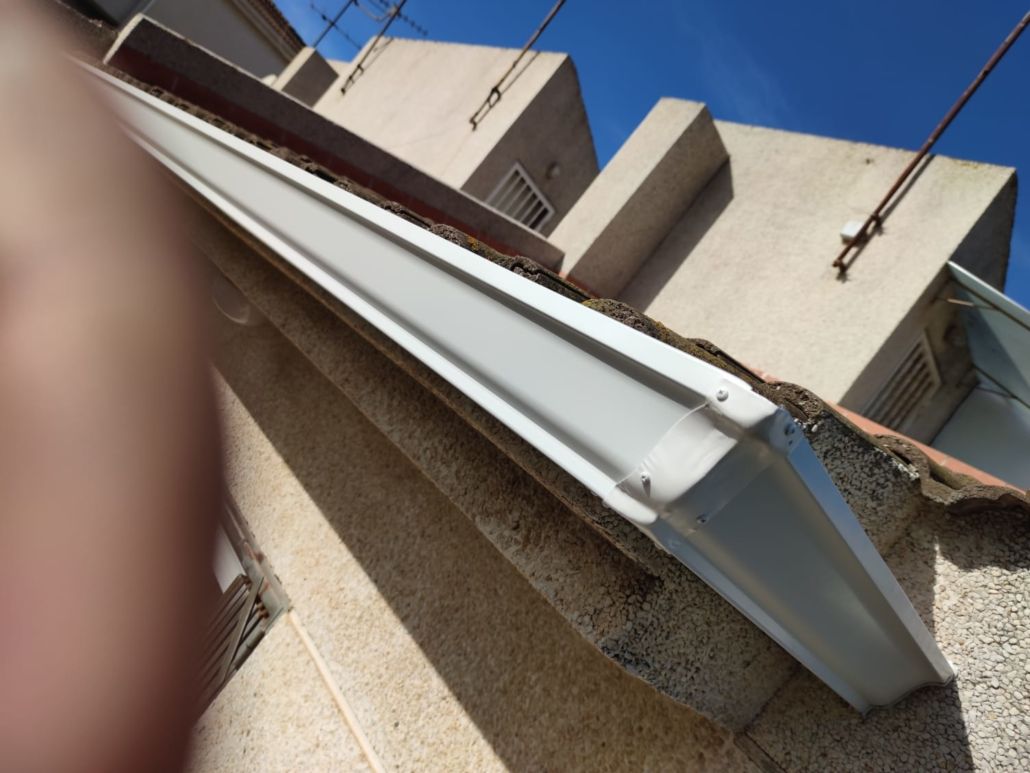 Vista desde un ángulo bajo de un canalón blanco instalado a lo largo del borde de un tejado de tejas, con un cielo azul despejado de fondo.