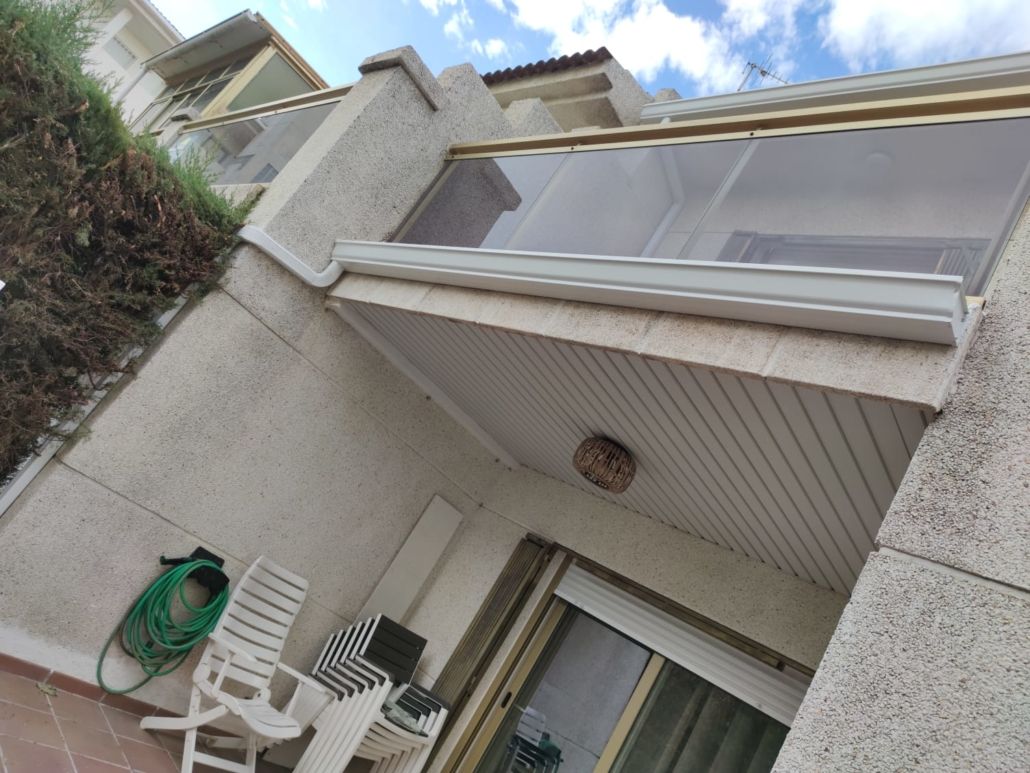 Vista desde un ángulo bajo del exterior de un edificio beige con un balcón cubierto, una manguera verde enrollada y sillas de plástico blancas.