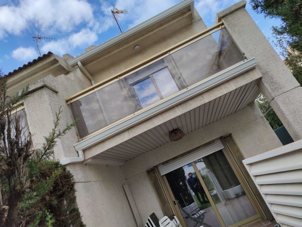 Una casa de dos plantas de color beige claro con un balcón con barandilla de cristal y puertas correderas de cristal en la planta baja, bajo un cielo azul.