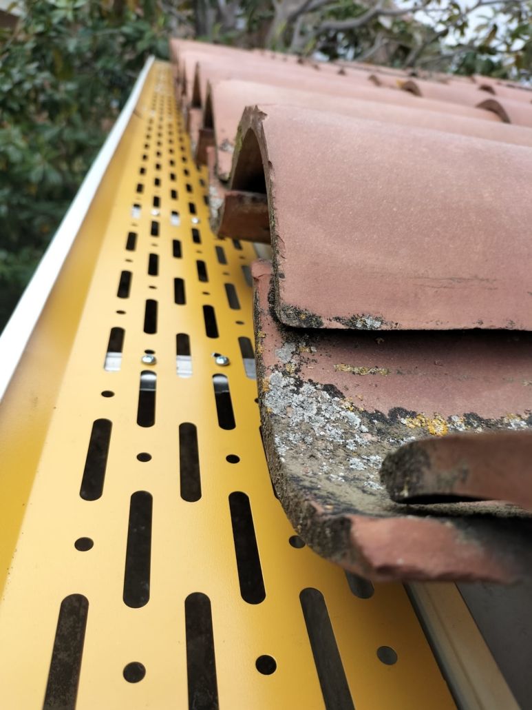 Un protector de canalón metálico ranurado de color amarillo instalado a lo largo del borde de un tejado con tejas de barro desgastadas por el tiempo.