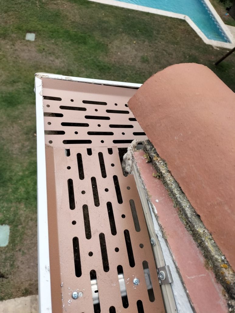 Vista en primer plano, desde arriba, de un panel de ventilación de techo marrón ranurado instalado junto a una teja curva de terracota.