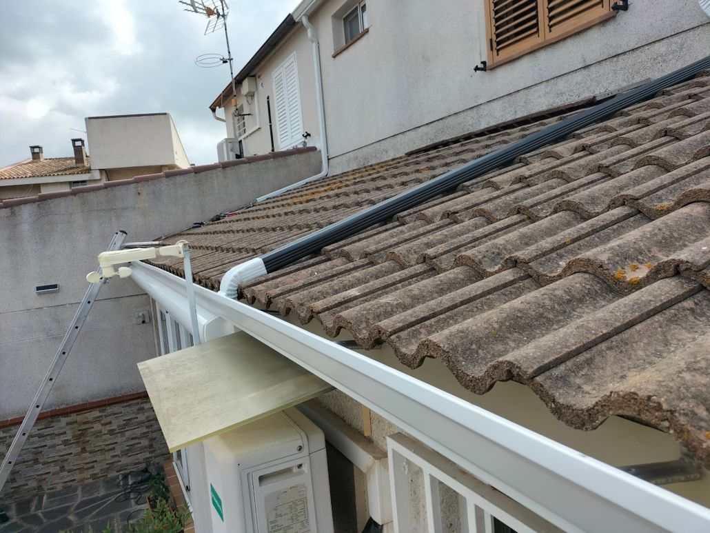 Una canaleta blanca está fijada al borde de un tejado de tejas, con una manguera negra que se extiende desde la canaleta a lo largo de la pendiente del tejado.