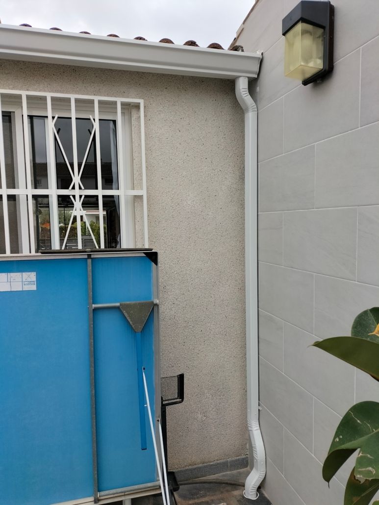 Un bajante blanco sobre una pared beige texturizada, junto a una pared de bloques de hormigón gris y una pantalla azul.