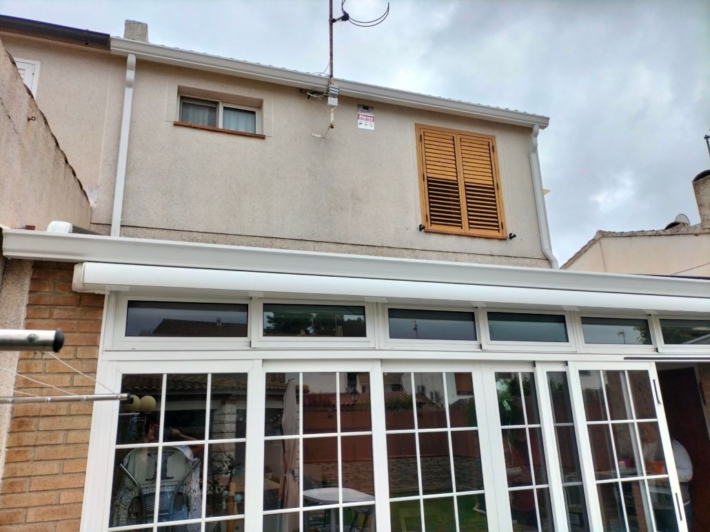 La parte trasera de una casa residencial con un invernadero blanco, una pequeña ventana en la parte superior y una contraventana de madera cerrada.