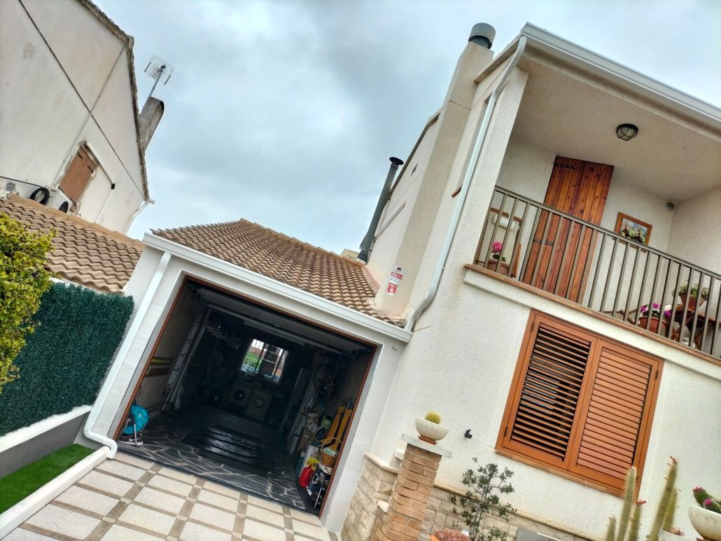 Una casa blanca de dos plantas con garaje con tejado de tejas y balcón con puerta y contraventanas de madera.