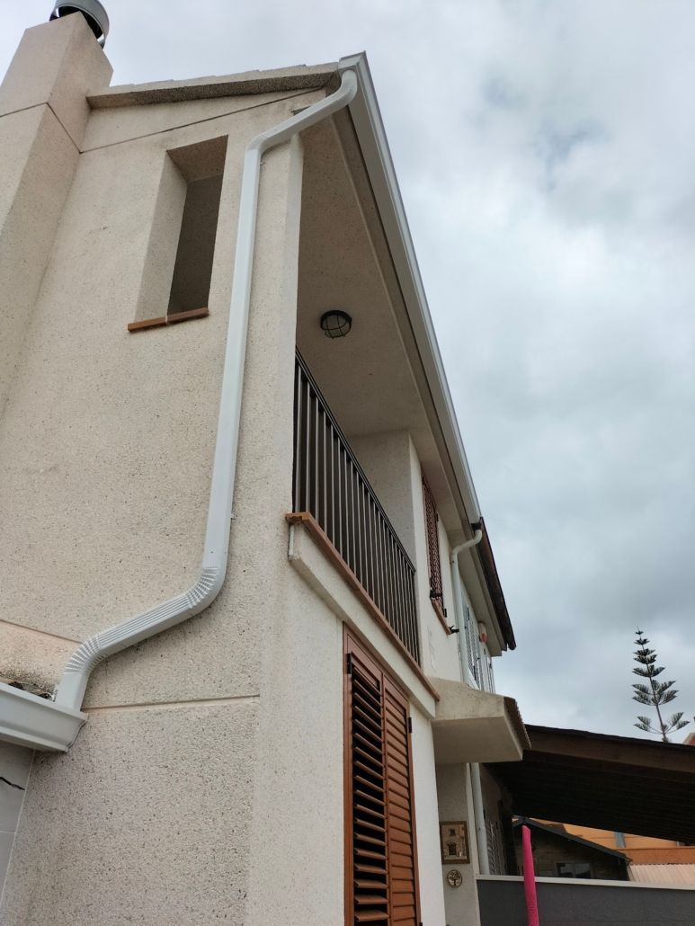 Vista desde un ángulo bajo de una casa beige de varias plantas con un pequeño balcón, contraventanas marrones y un sistema de canalones exteriores.