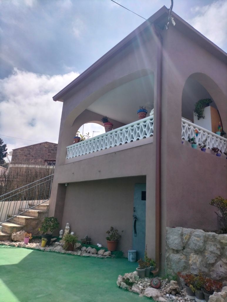 Un edificio de dos plantas de color gris topo con un balcón arqueado con barandilla blanca y una puerta a nivel de la calle que da a un patio verde.