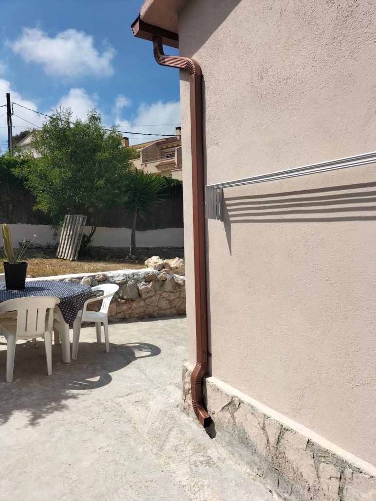 Un bajante de metal marrón recorre verticalmente una pared exterior de estuco beige junto a un patio con muebles de exterior.