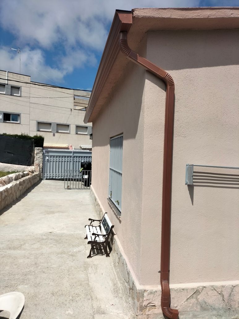 Un bajante marrón en una pared exterior de color claro, junto a un pequeño banco en un sendero pavimentado cerca de un edificio de varias plantas.