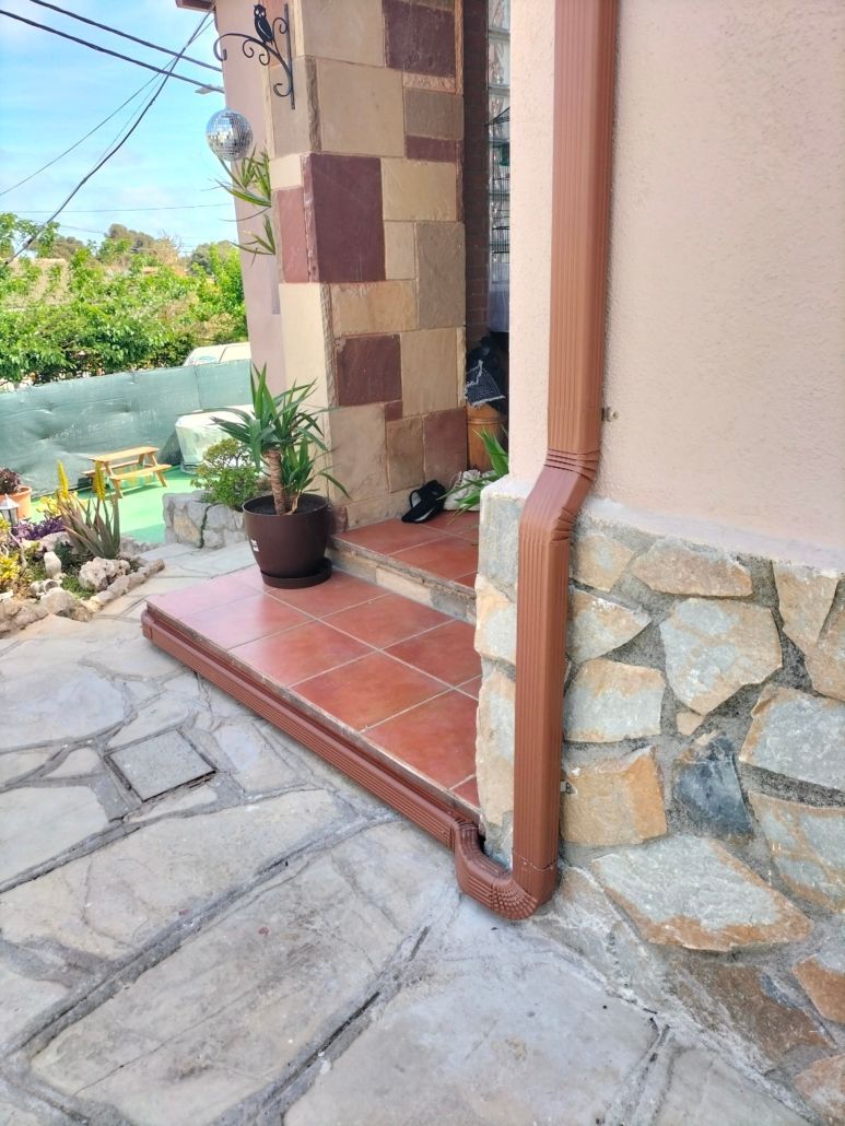 Una entrada con porche delantero, un escalón de baldosas de terracota, un pilar de piedra, una planta verde en maceta y un bajante de metal marrón.