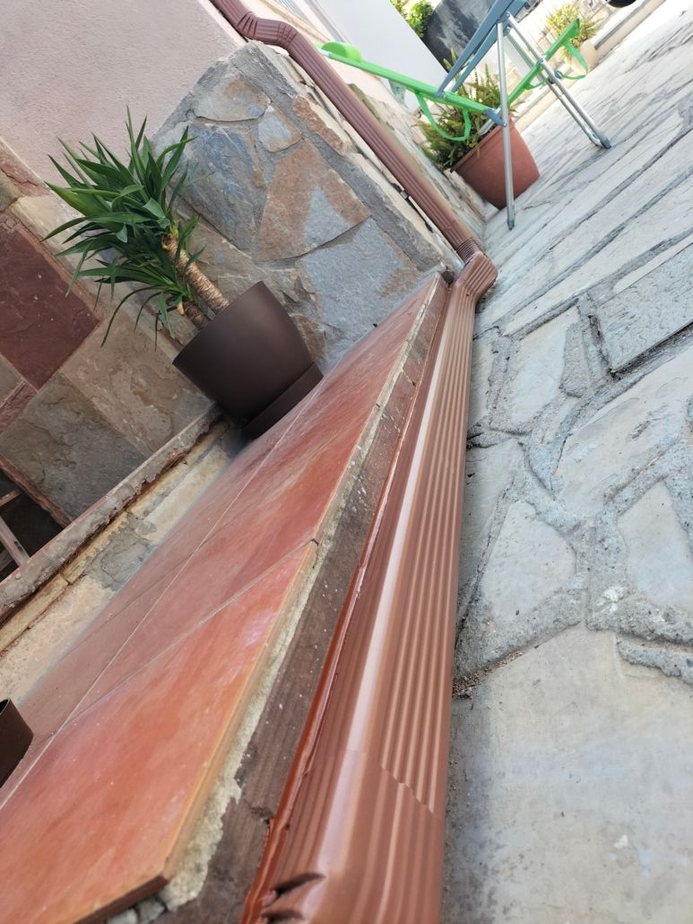 Vista de cerca de una canaleta corrugada marrón sobre un patio de piedra, junto a un escalón de baldosas con plantas en macetas.
