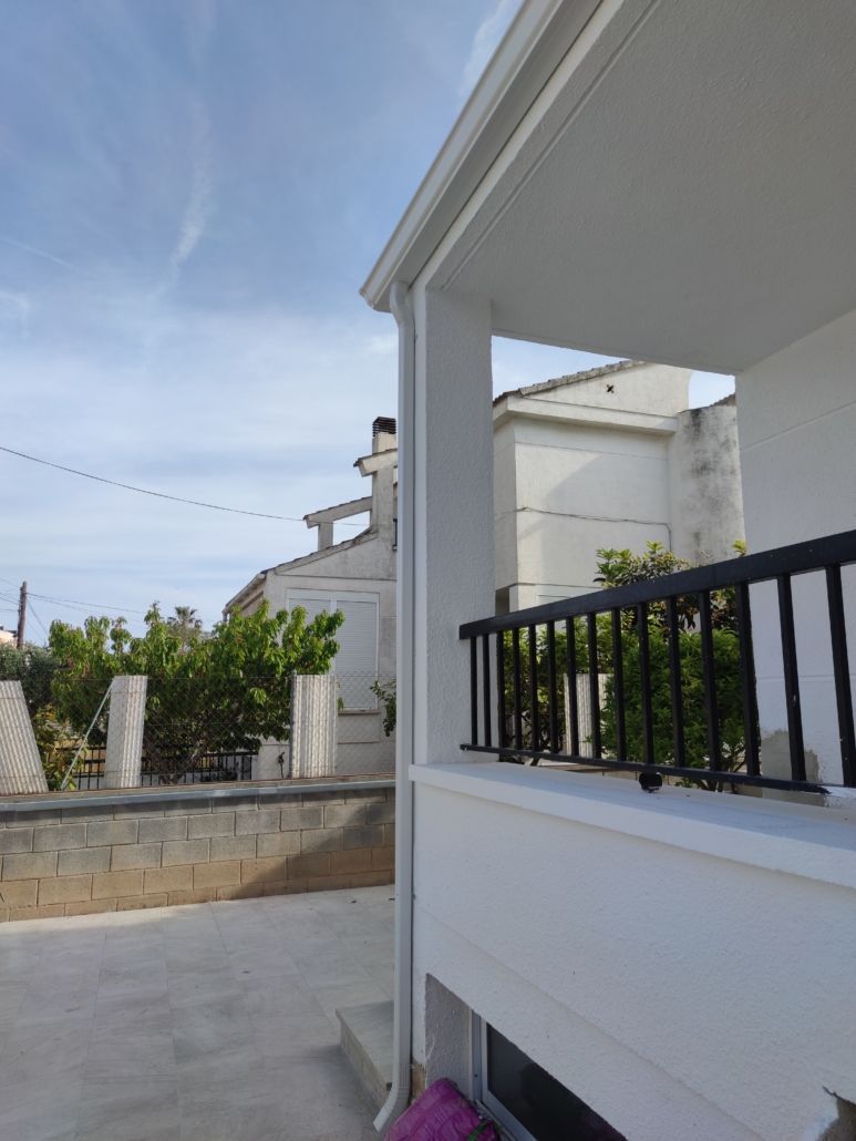 Vista de una esquina de un edificio blanco con balcón, barandilla negra y patio con baldosas bajo un cielo brillante y soleado.