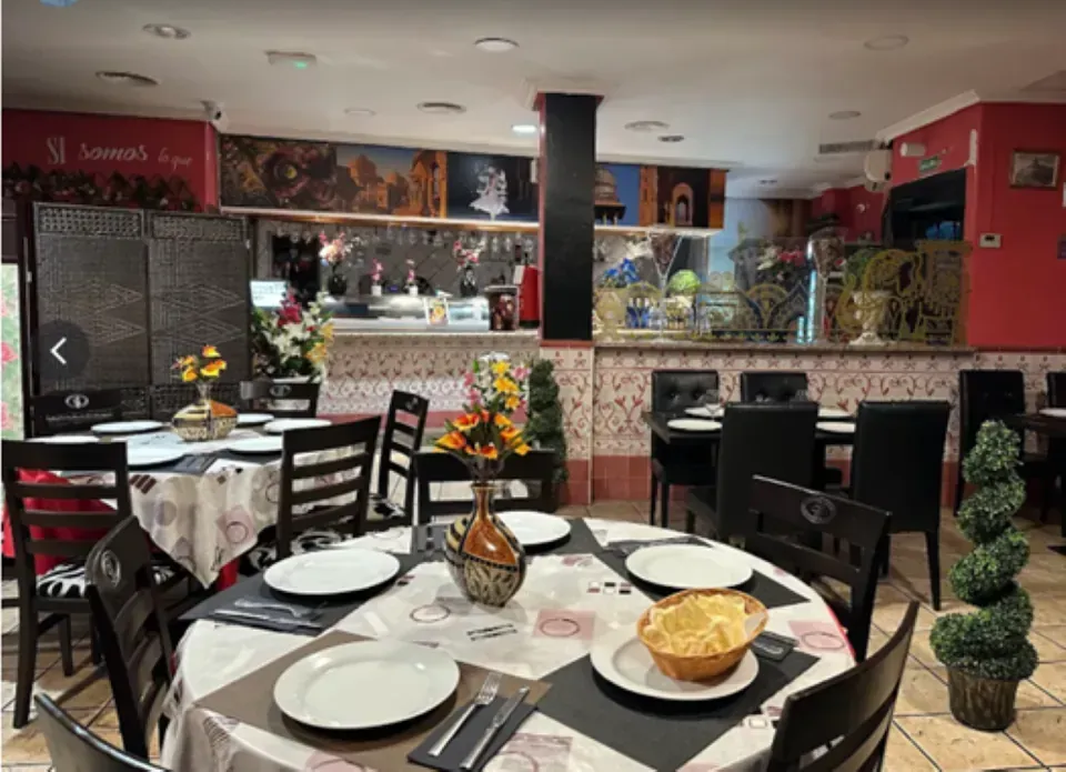Interior de restaurante con mesas preparadas para cenar; flores decorativas y obras de arte; bar al fondo.