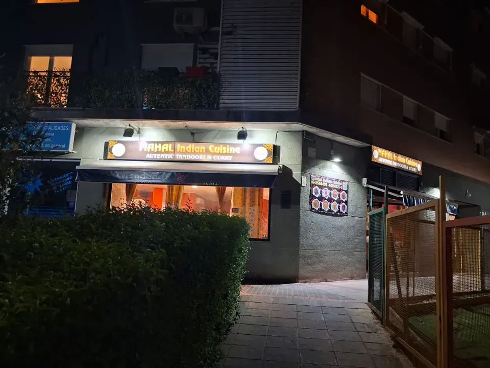 Un restaurante indio de noche con letrero y entrada iluminados. El edificio es gris y hay una pequeña pasarela.