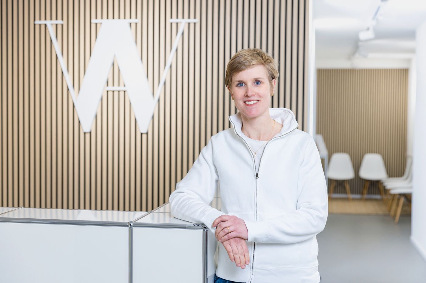 Frau in weißem Kapuzenpullover steht an einem modernen Empfangstresen vor einer Holzwand mit einem „W“-Logo.