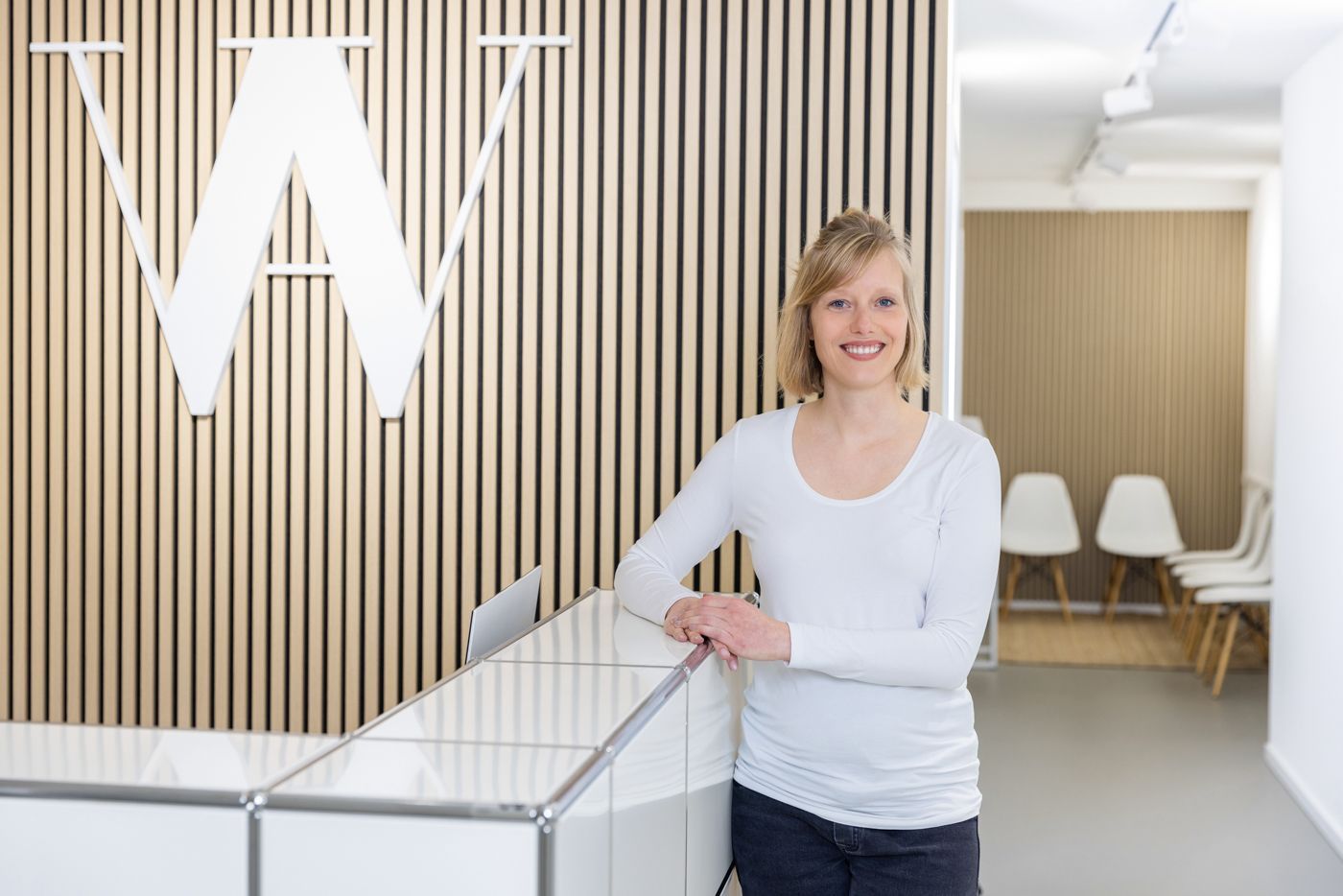 Frau an der Rezeption lächelt, weißes Oberteil, holzgetäfelte Wand mit „W“-Logo, Stühle sichtbar.