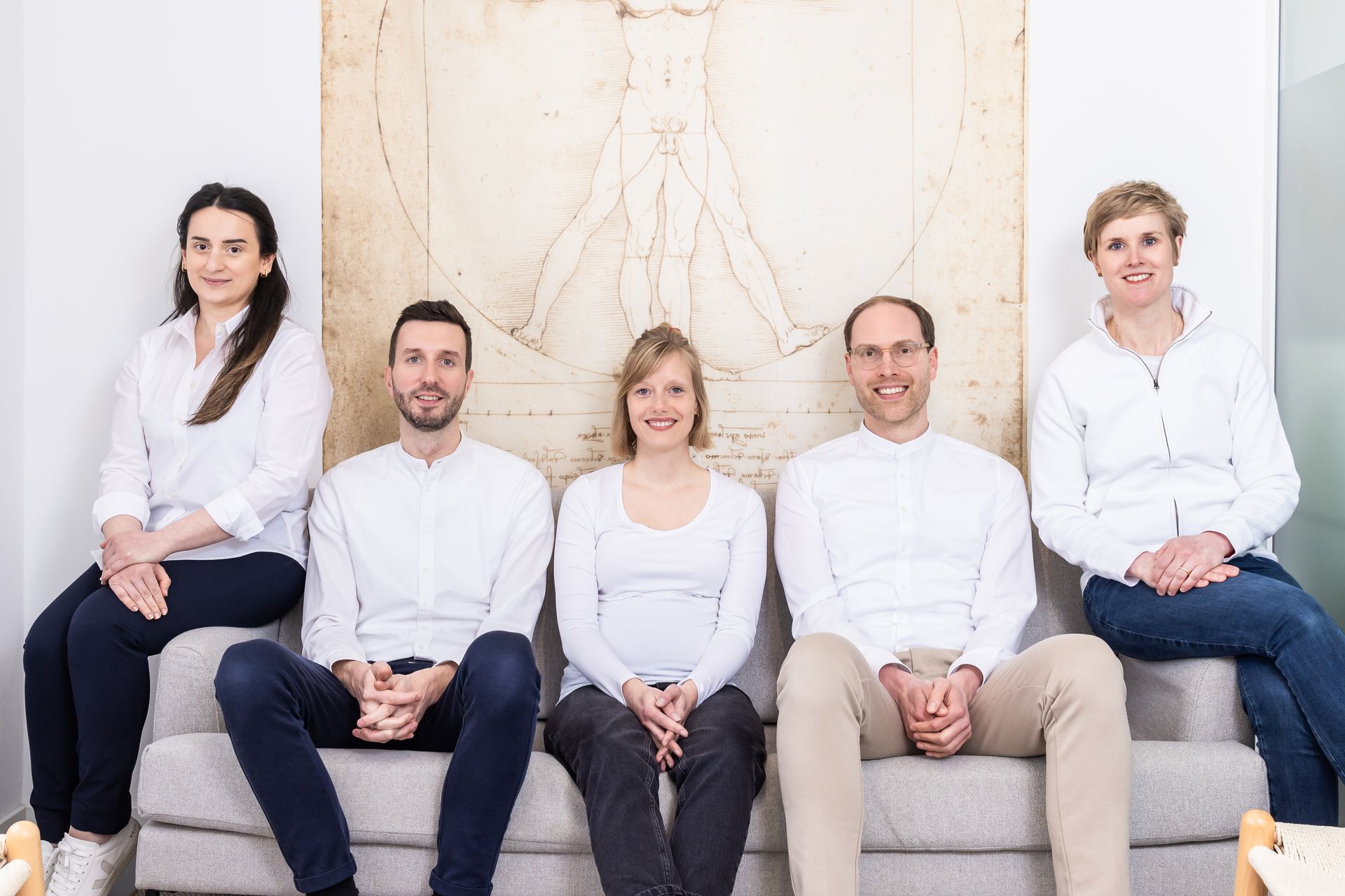 Fünf Personen in weißen Hemden und Jeans/Hosen sitzen auf einem grauen Sofa vor einer Wand mit einer Zeichnung.