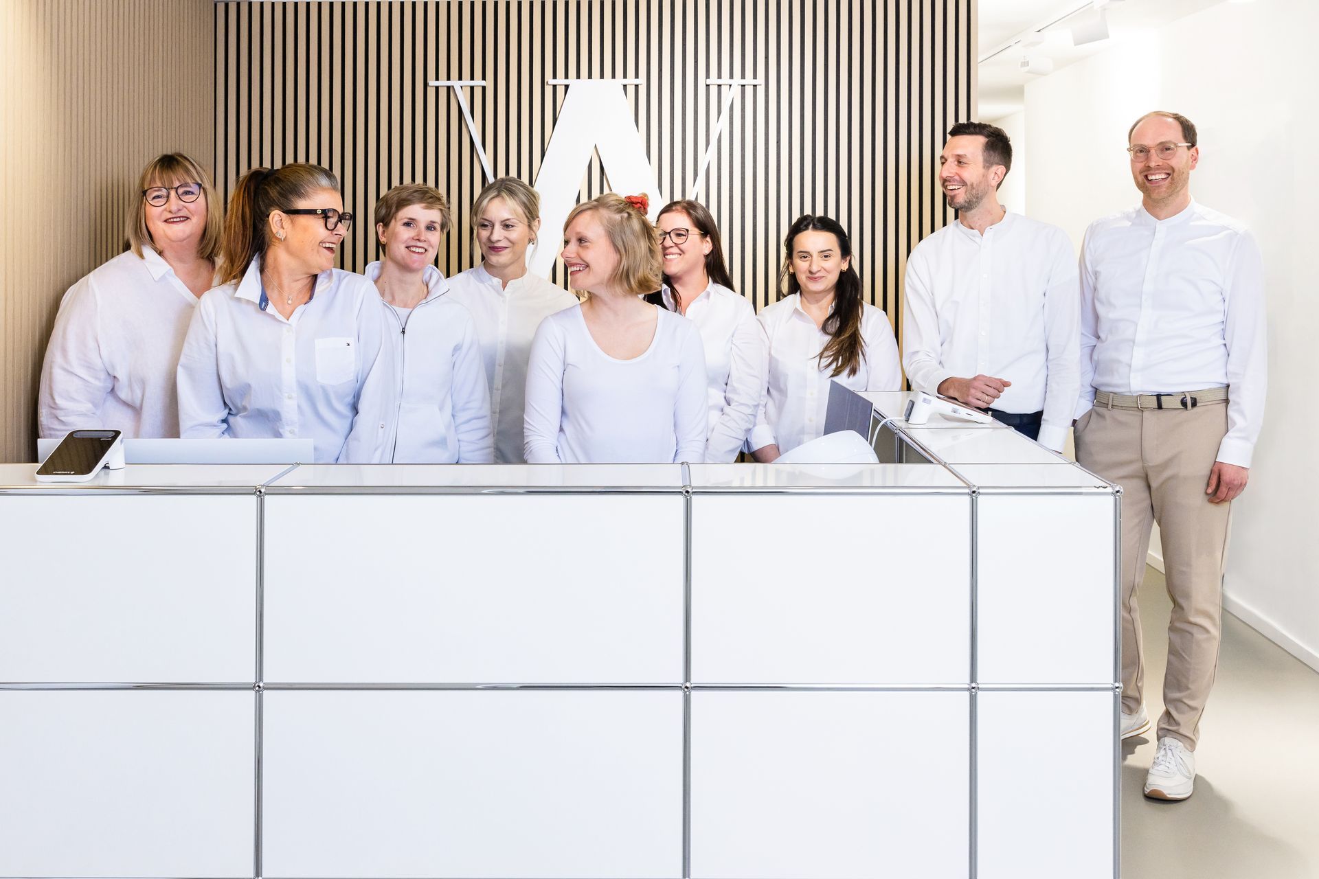 Gruppe von Menschen in weißen Hemden hinter einem weißen Empfangstresen mit einem Logo in einem hellen Büro.