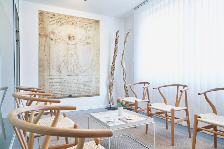 Ein Wartezimmer mit Holzstühlen, einem kleinen Tisch und einer gerahmten anatomischen Zeichnung.
