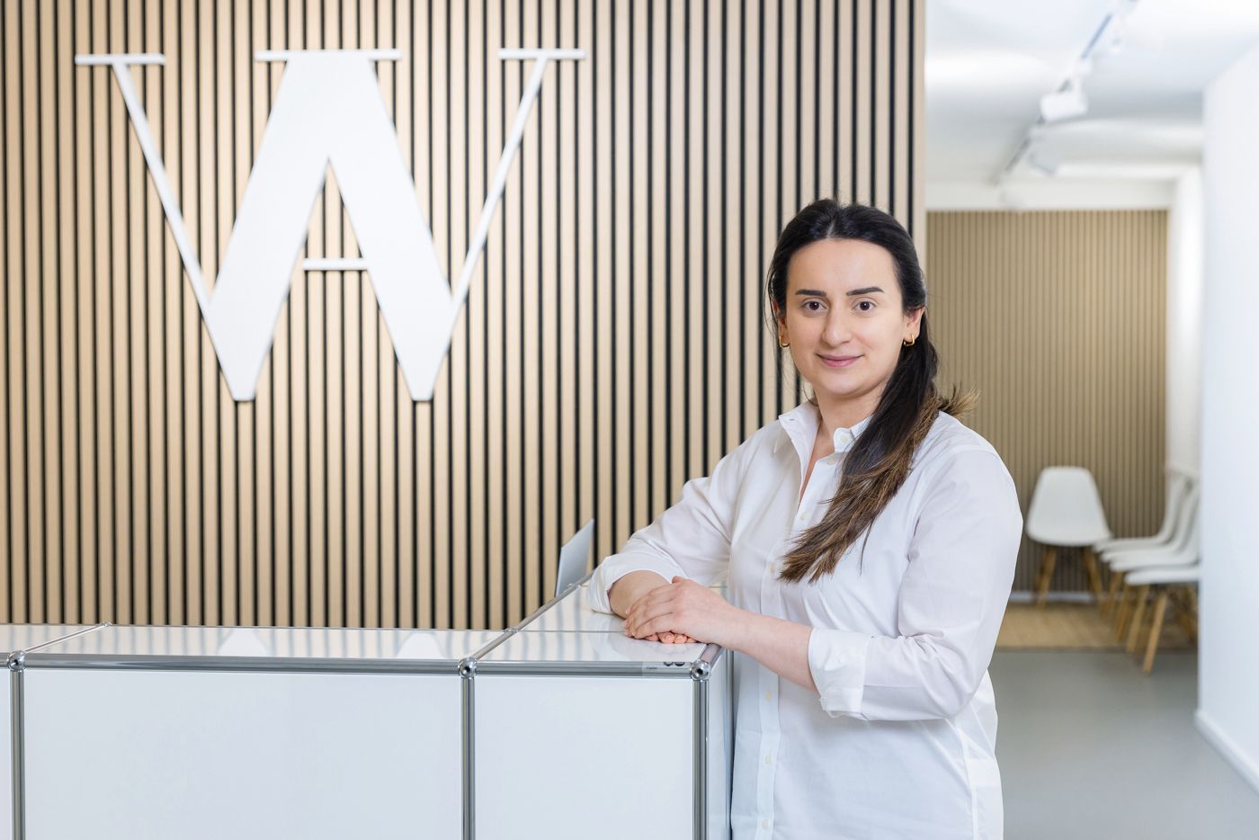 Frau an einem Empfangstresen mit „W“-Logo, weißes Hemd, Holzwand.