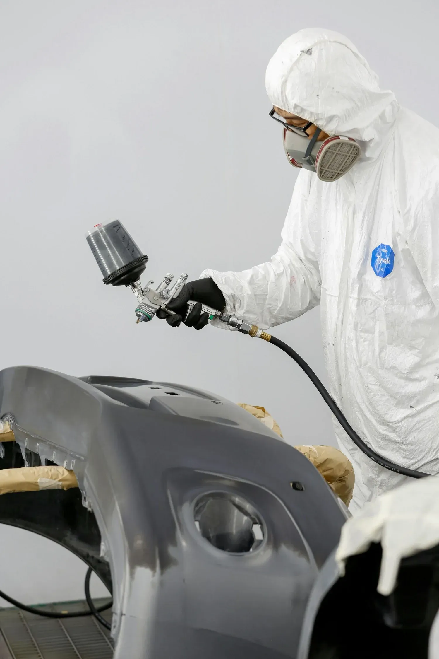 Persona con traje de protección rociando pintura sobre el parachoques de un automóvil.