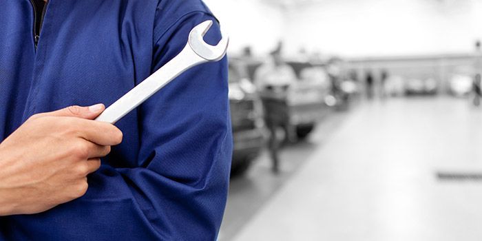 Un mécanicien en combinaison bleue tient une clé à molette sur un fond flou de garage.