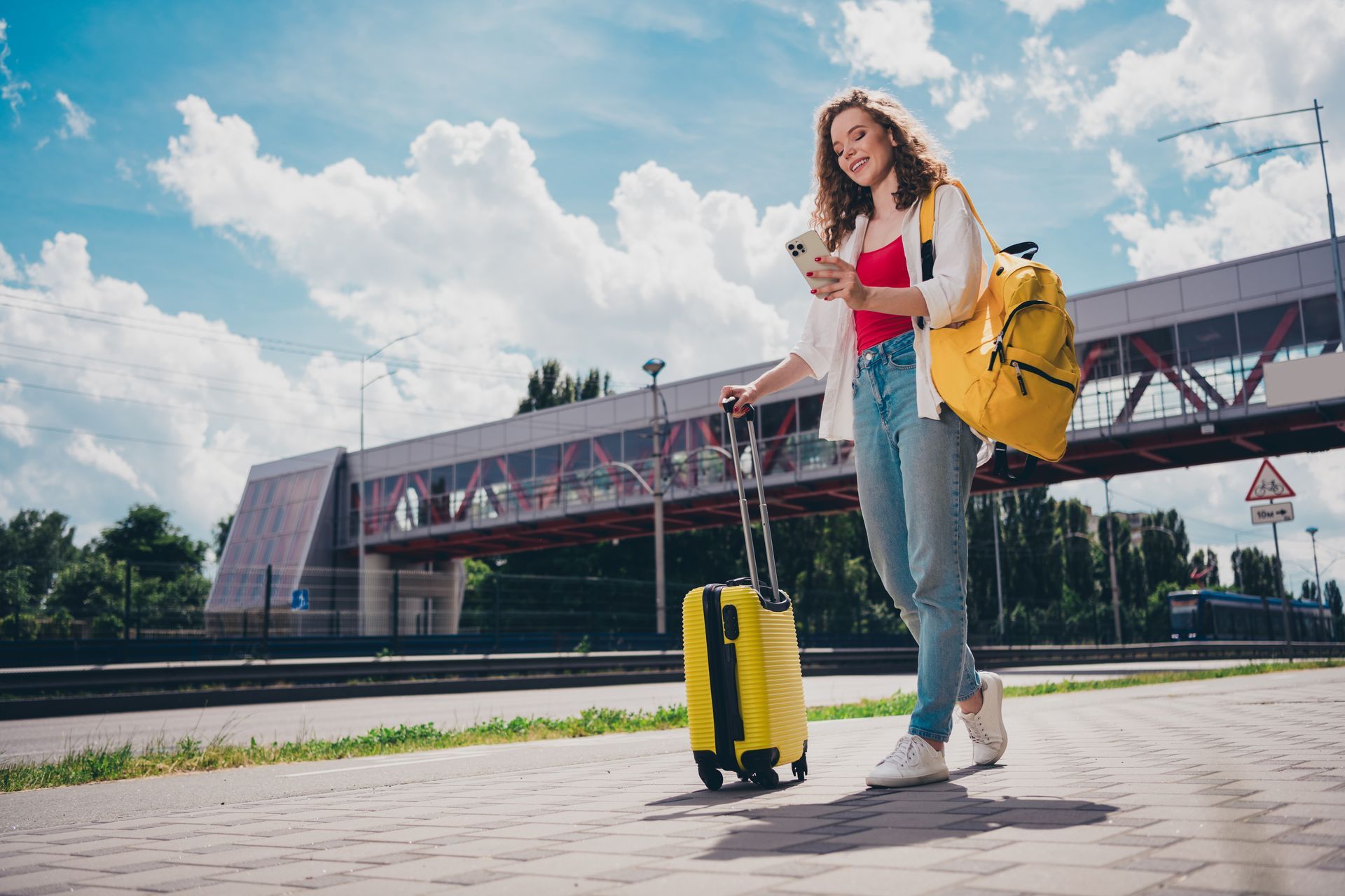 Femme avec valises et sac à dos jaunes, utilisant son téléphone près d'un pont moderne.