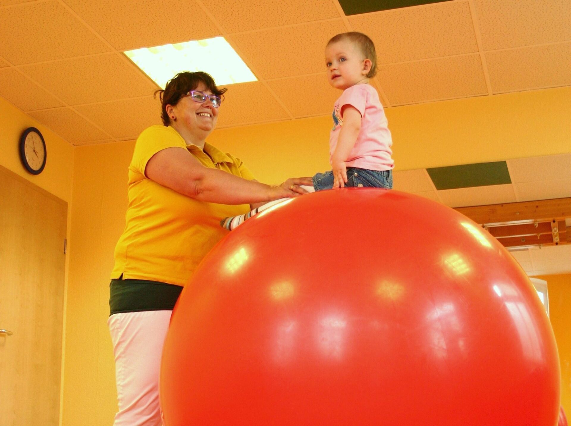 Eine Ergotherapeutin betreut ein kleines Kind, sitzend auf einem roten Gummiball