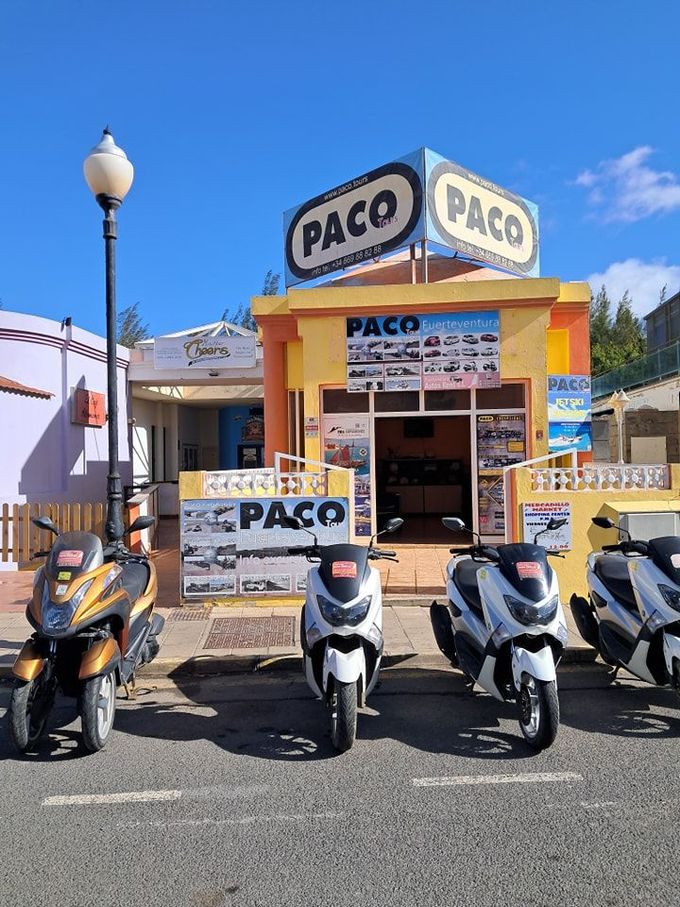 Pequeña tienda PACO con scooters aparcados delante en una calle soleada.