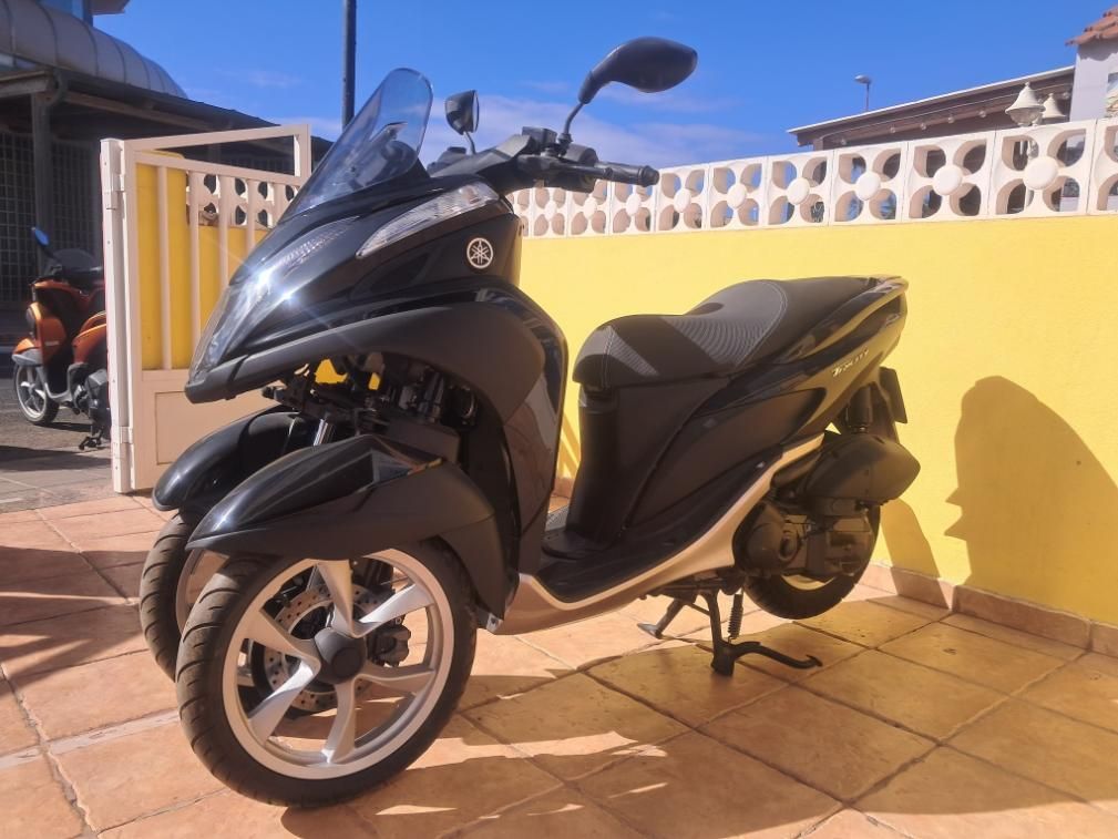 Una moto negra aparcada en un soleado patio embaldosado junto a una pared amarilla y una valla blanca.