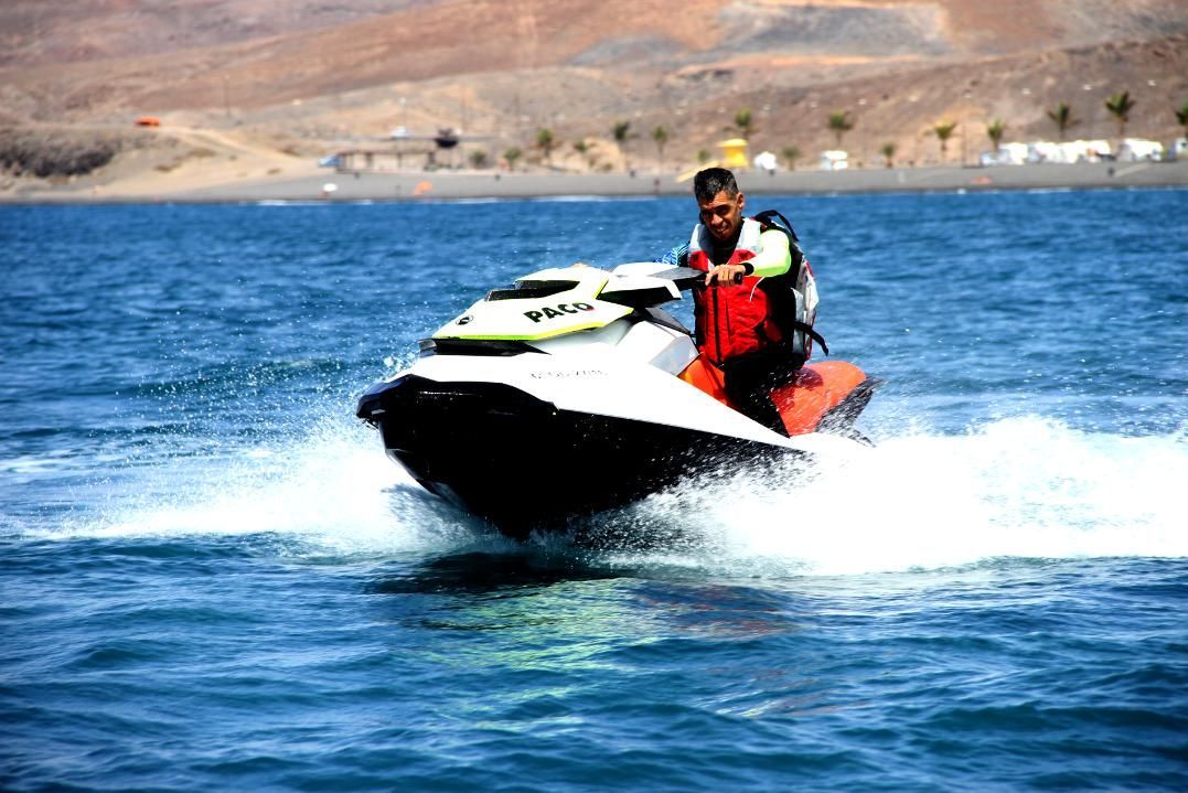 Una persona monta una moto acuática blanca en agua azul, con una costa rocosa al fondo.