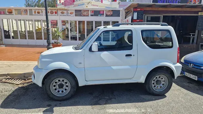 Un SUV compacto blanco estacionado en una calle frente a unas tiendas, con un coche azul al lado.